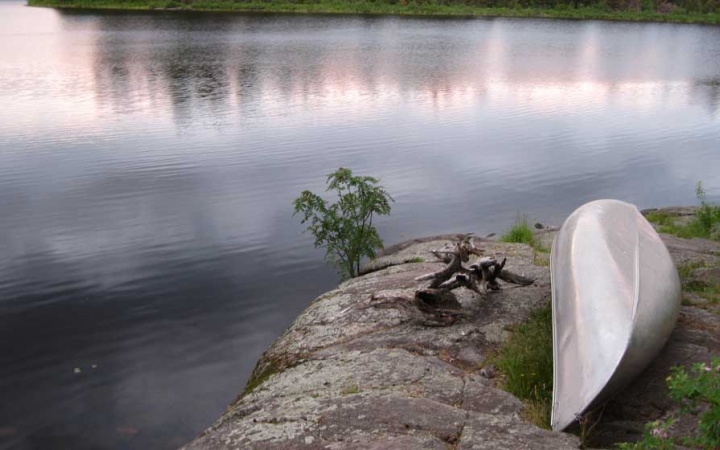 A canoe rests upside down on a rocky shore beside a very calm body of water. 